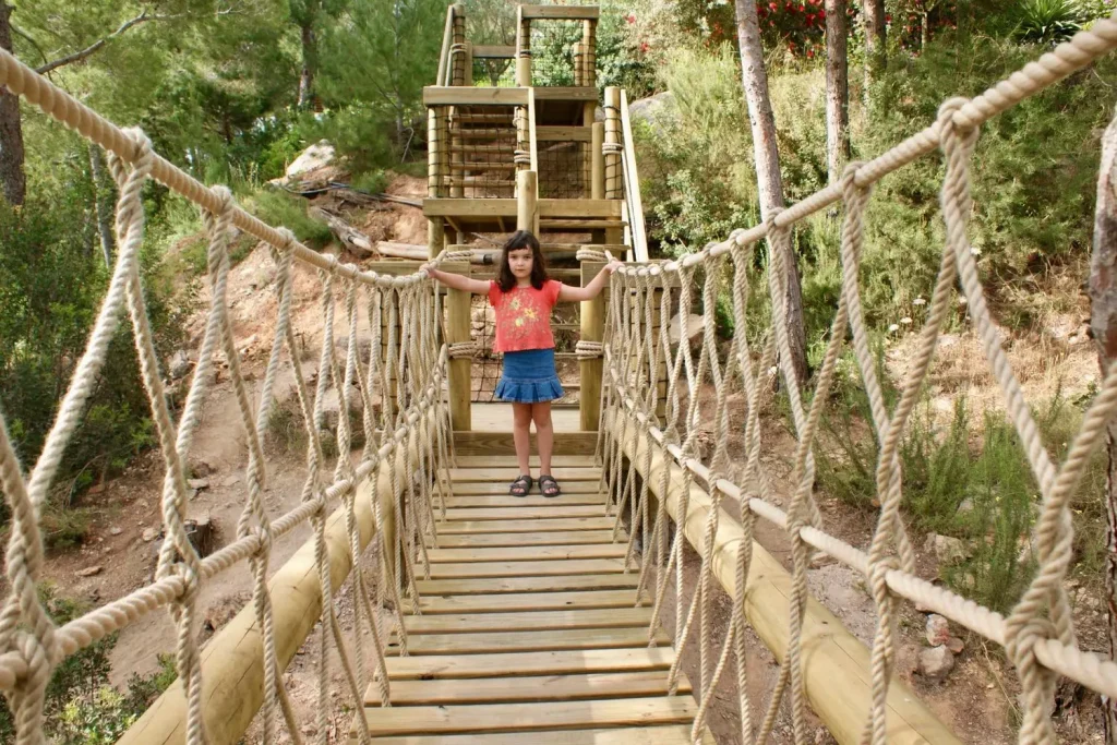 Girl child standing on a playful Treehouse Rope Bridge set into a Mediterranean family garden with steps and stairs in the background.