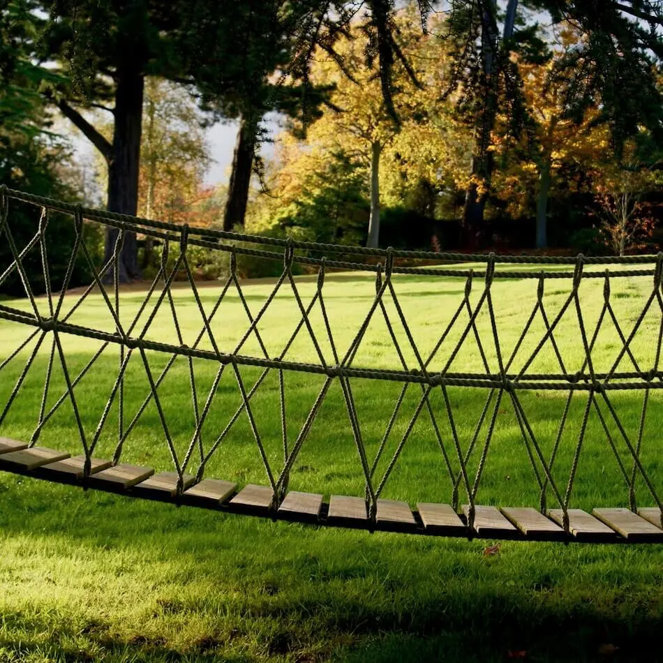 View of Rope Bridge from side looking at rope work with lawn and trees behind.