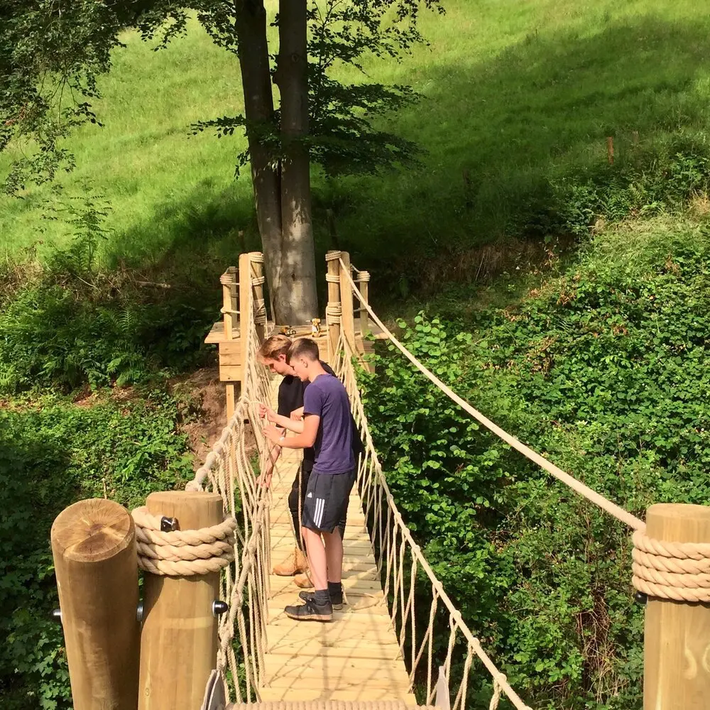 Men working on family garden suspended Rope Bridge across a river ravine with trees and fields in the background.