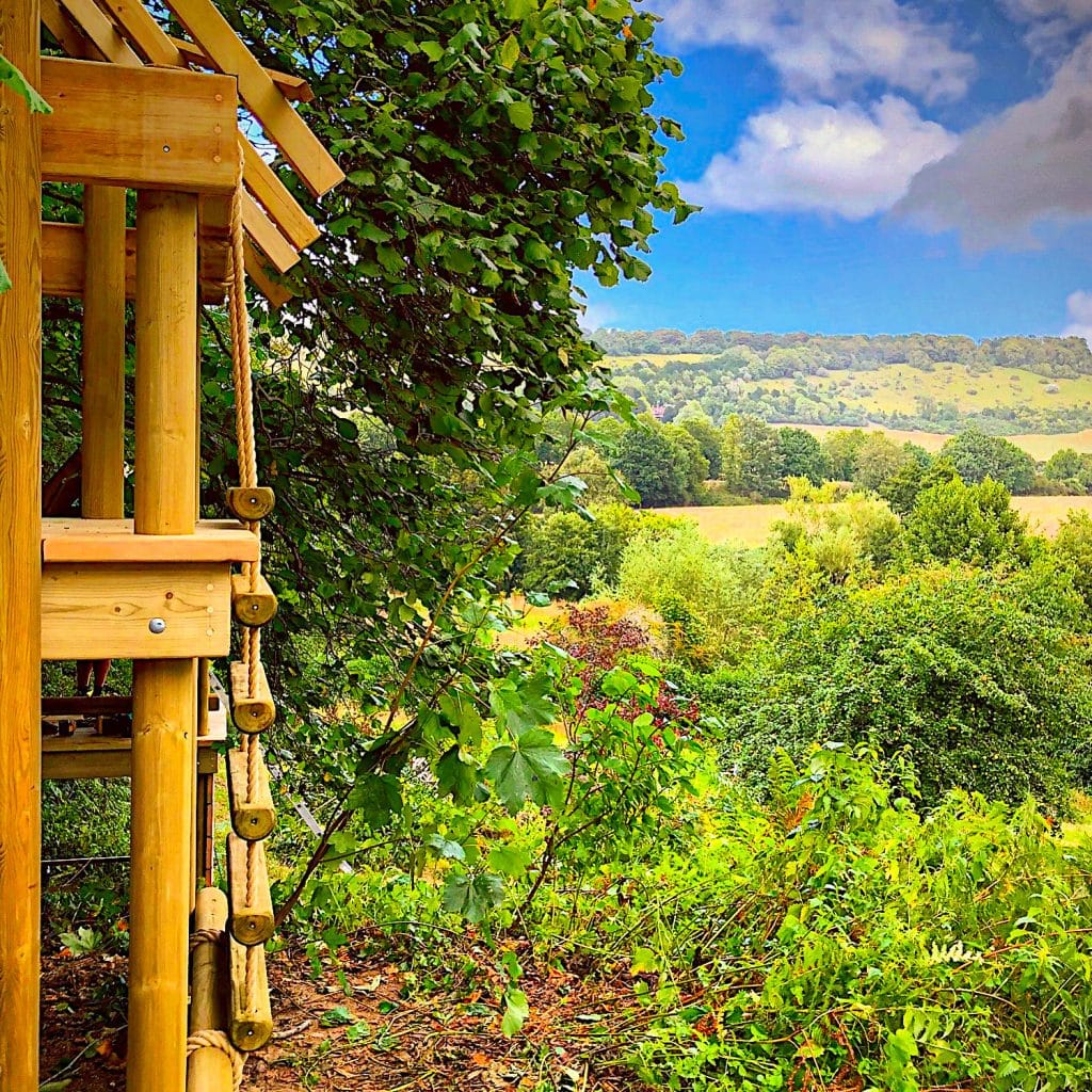 The corner of a wooden structure with rope details overlooks a scenic landscape, offering a natural observation point amidst lush foliage.