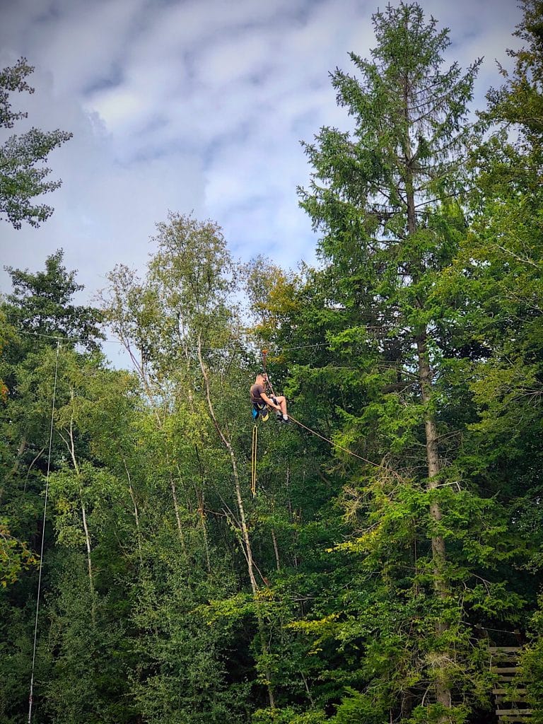 A man suspended by rope between two high trees as he works on the installation on an aerial park in the UK. He is suspended by a caribena and harness and rope.
