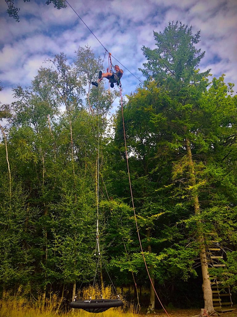 A person suspended high in the air between two giant fir trees. They are hoisting up a nest swing on a harness and rope. A person in mid-action on a high ropes course in a forest setting, showcasing the energy and skill involved in navigating aerial challenges