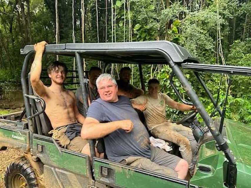 A team of 5 master rope bridge builders at Treehouse Life stop in their open jeep in the jungle to pose for a photo. They are working on a project on Kauai Island in Hawaii building 4 Rope Bridges. The Jeep is stopped on muddy rough terrain and surrounded by jungle trees and long vines. the Treehouse Life team look happy and are smiling at the camera.