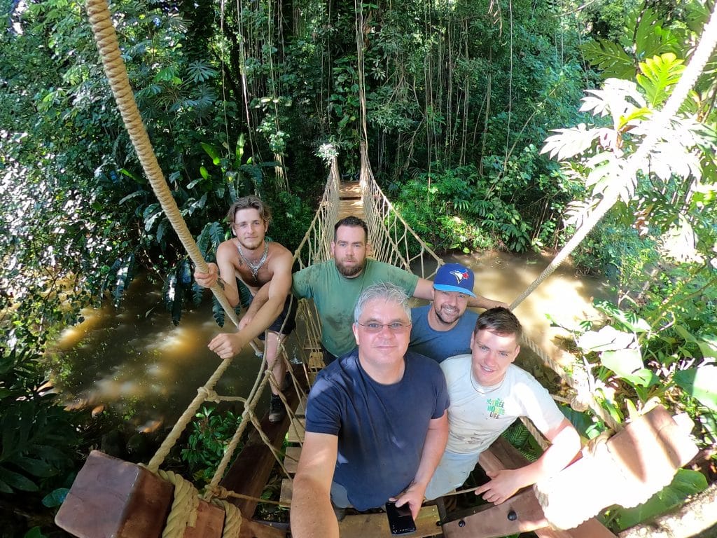 Paul, Charlie and the wider team at Treehouse Life posing for a group photo. The group stand at the end of a rope bridge in the jungle landscape of Kauai Island in Hawaii.
